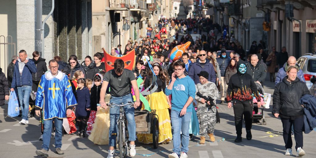 Carnevale a Borgomanero. Corso Mazzini pieno di persone in maschera, capitanate da un piccolo carro carnevalesco trainato da una bici